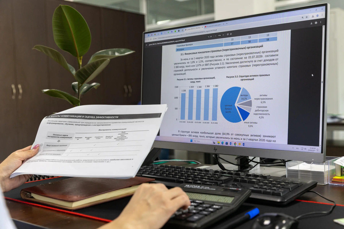 A person reviews financial charts and printed reports at an office computer.