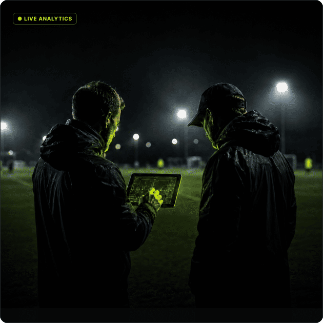 Two coaches reviewing live squad analytics on a tablet beside a floodlit pitch