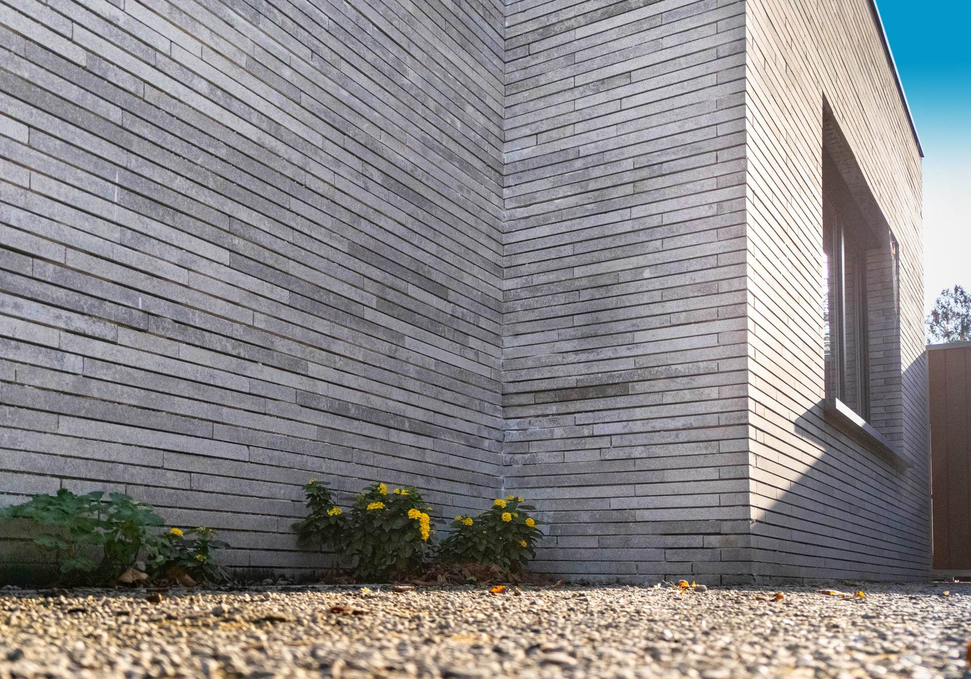 Hoek van de gevel van vlak boven de grond gefotografeerd, met een grindstrook en bloeiende beplanting aan de voet van het metselwerk