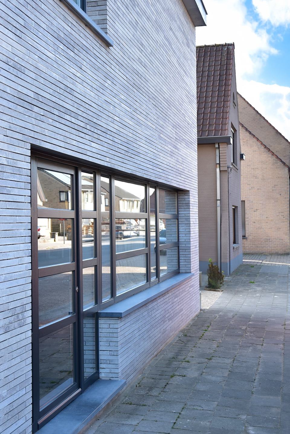 Straatzijde van de woning met een grote raampartij met zwarte profielen op het gelijkvloers, een voetpad in klinkers en een oudere buurwoning erachter