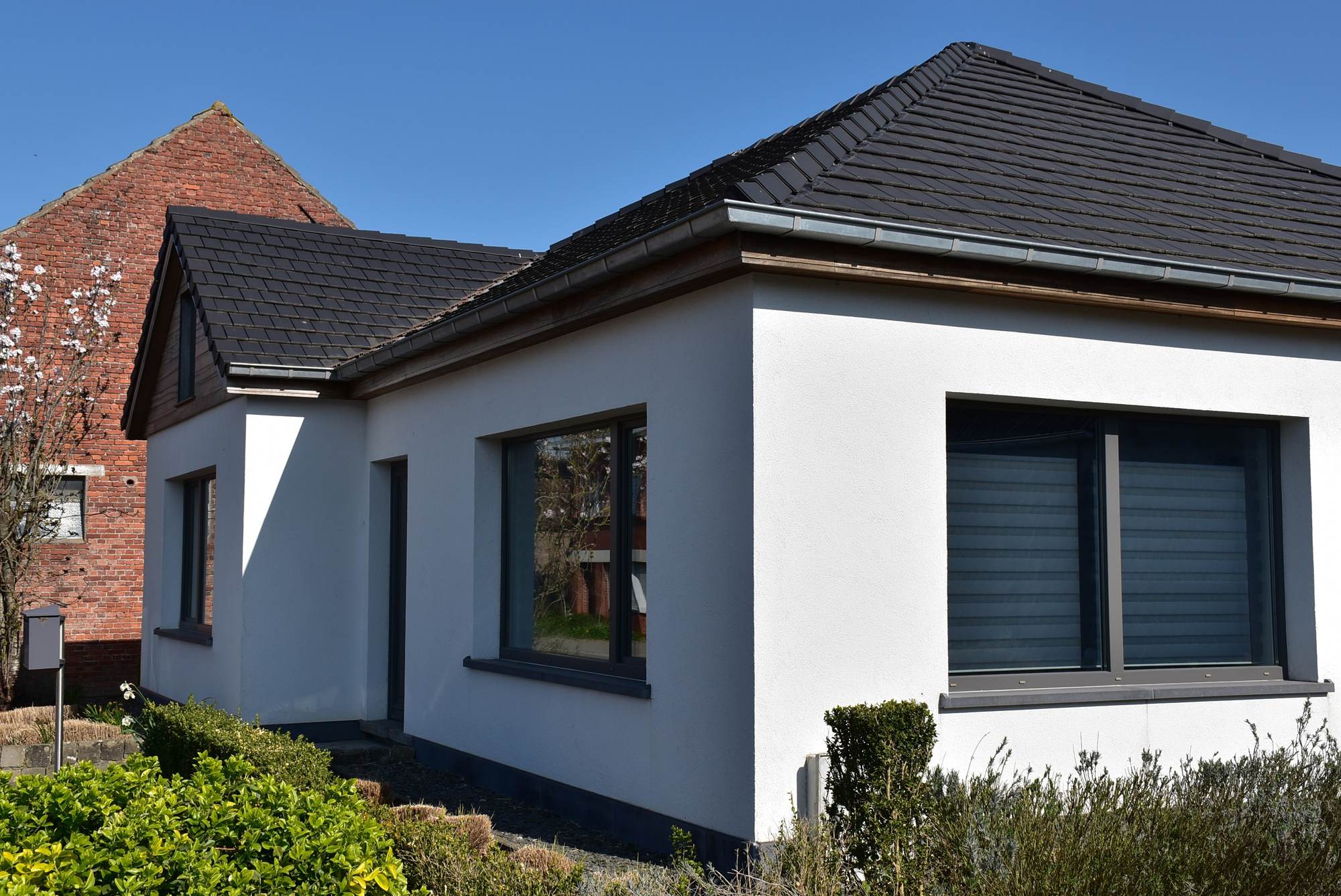 Hoek van een woning in Sint-Laureins met een gevel in lichte crepi onder een schilddak in matte antracietkleurige pannen, met een houten dakrand en een oude bakstenen puntgevel erachter