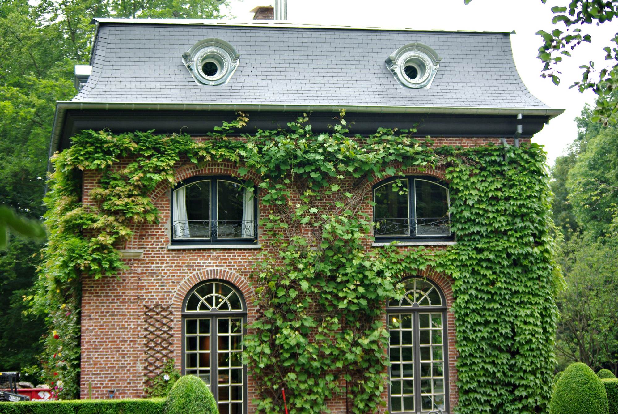 Herenhuis met een mansardedak in leien en twee ronde dakkapellen met een gerolde kap en een sierlijk profiel in grijs metaal, boven een bakstenen gevel begroeid met wingerd