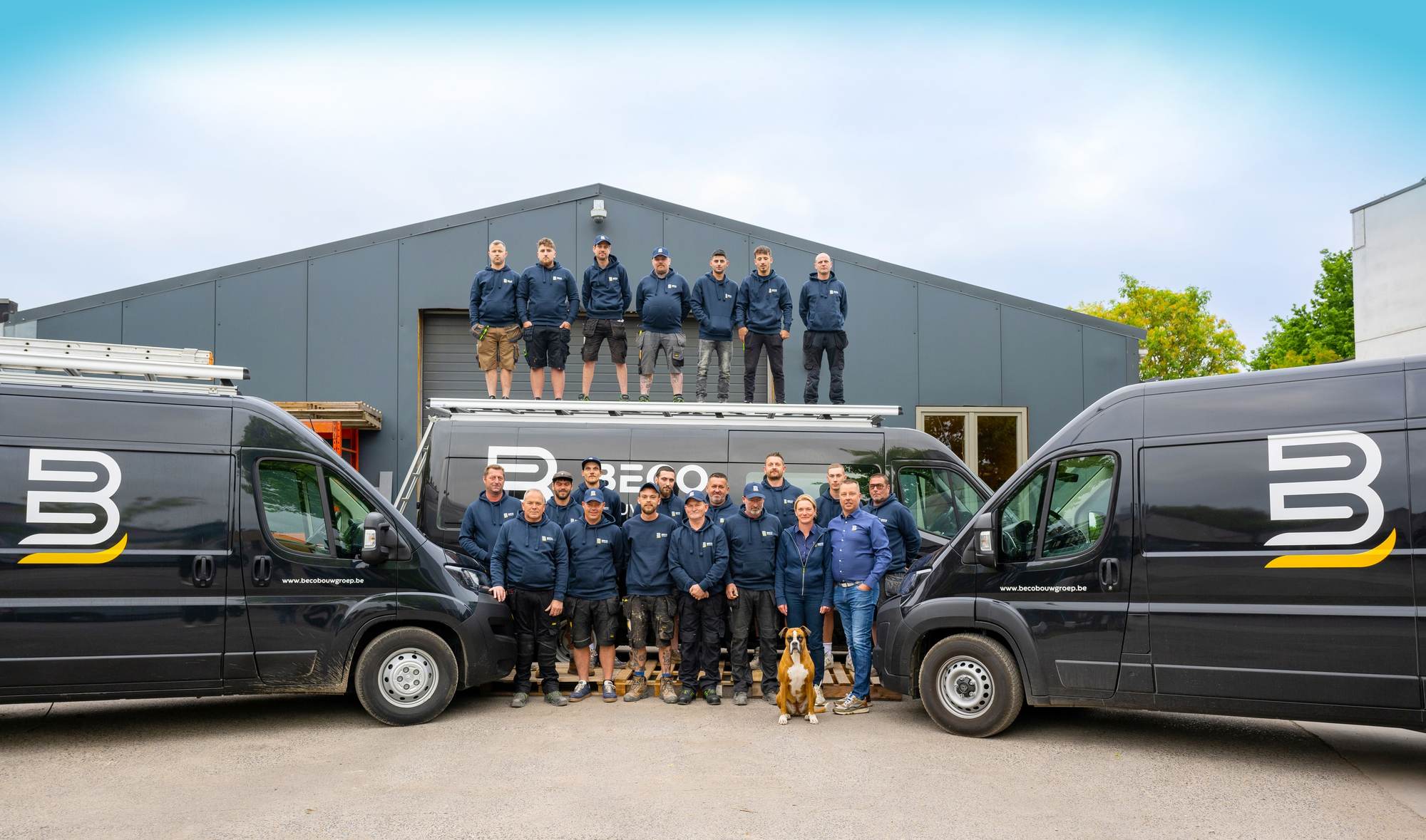 De volledige ploeg van Beco Bouwgroep staat tussen twee bedrijfsbestelwagens voor de loods, met een deel van de ploeg op het dak van een derde bestelwagen