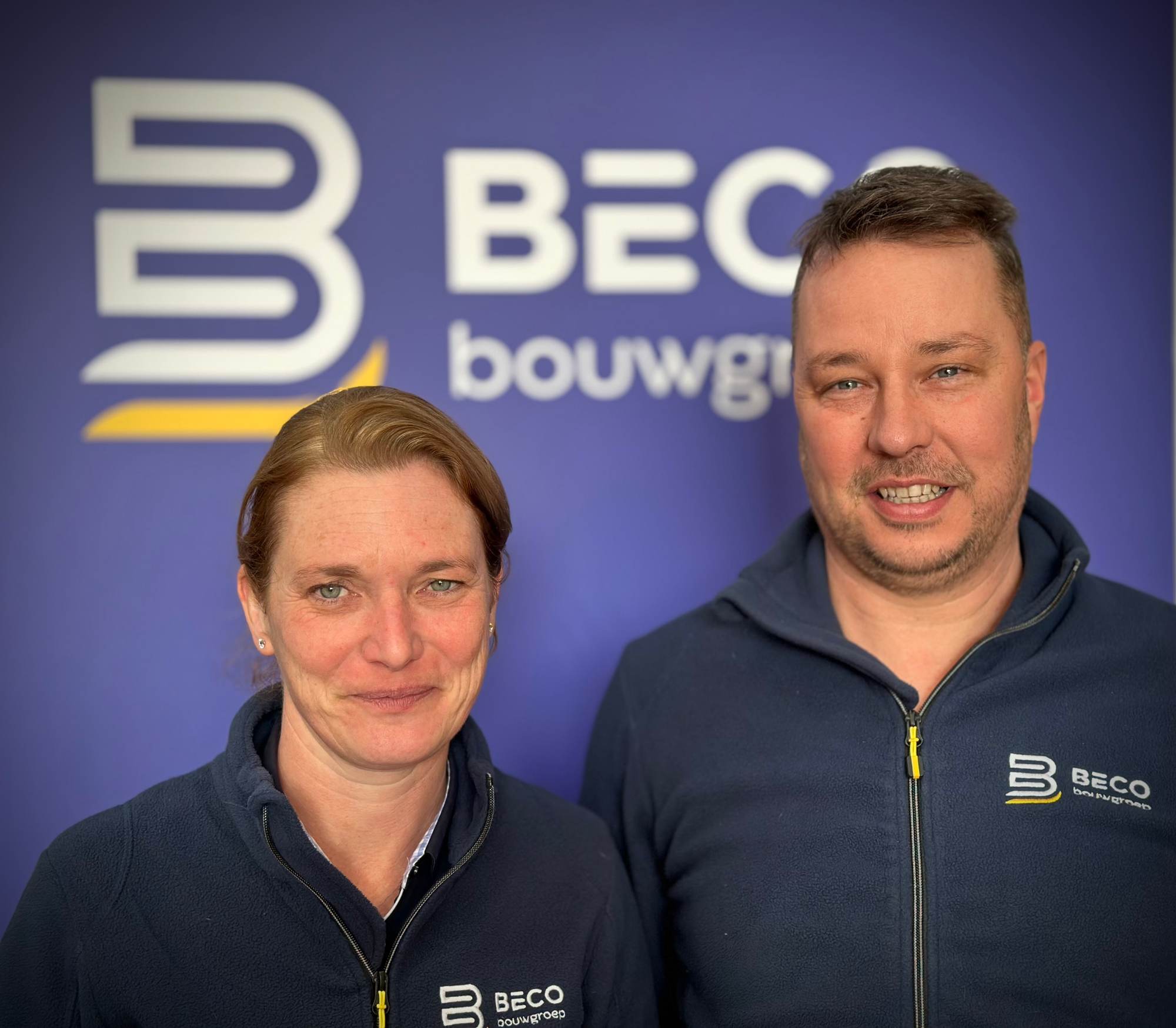 Annelies Cornilly en Benny Vanhaverbeke, de twee vennoten van Beco Bouwgroep, in bedrijfskledij voor een wand met het logo