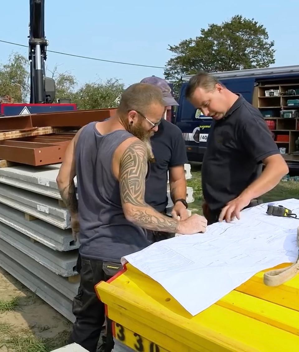 Drie mensen van de ploeg buigen zich op een werf over een bouwplan, met welfsels, stalen balken en de bestelwagen op de achtergrond