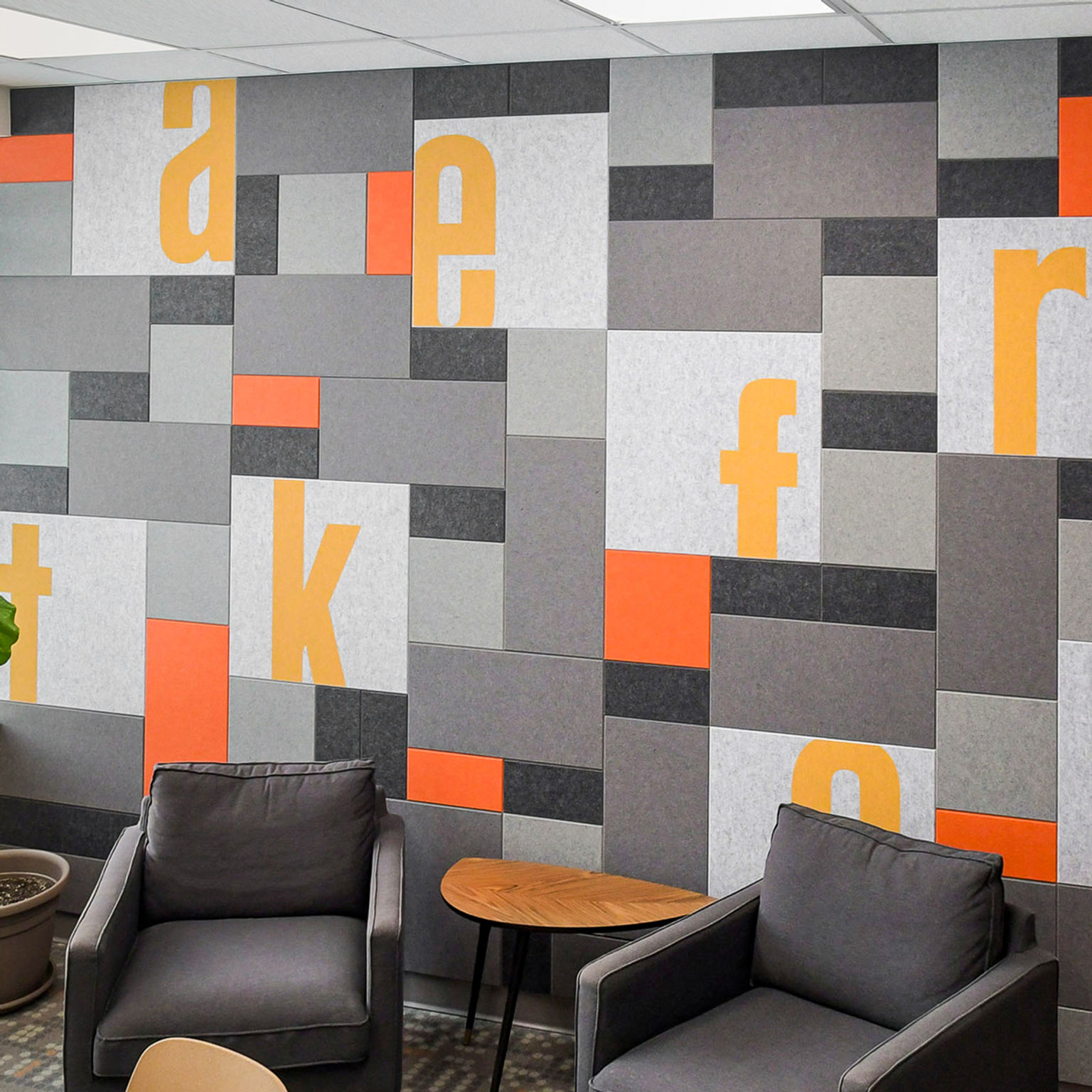 An office wall with an acoustic wall tile installation made up of rectangular tiles of various sizes and shades of gray accented by custom printing and bright orange tiles.