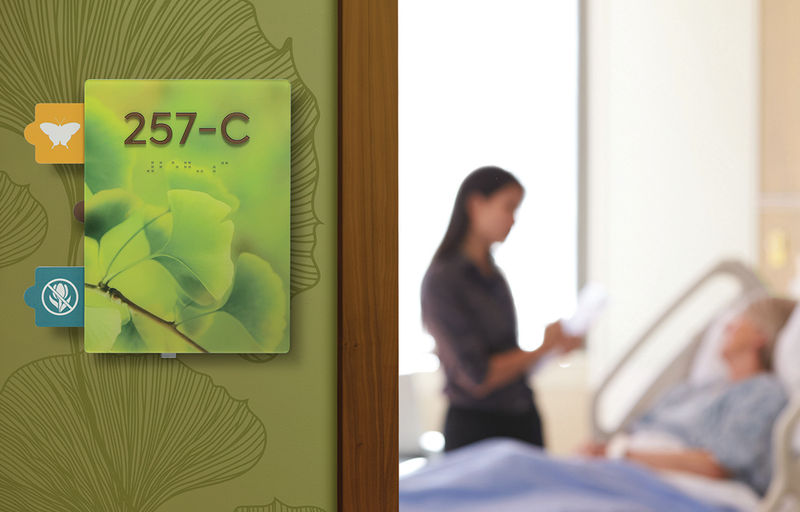 A three-tab ADA-compliant nurse alert patient room sign is positioned next to the open door; a nurse and patient can be seen in the room.