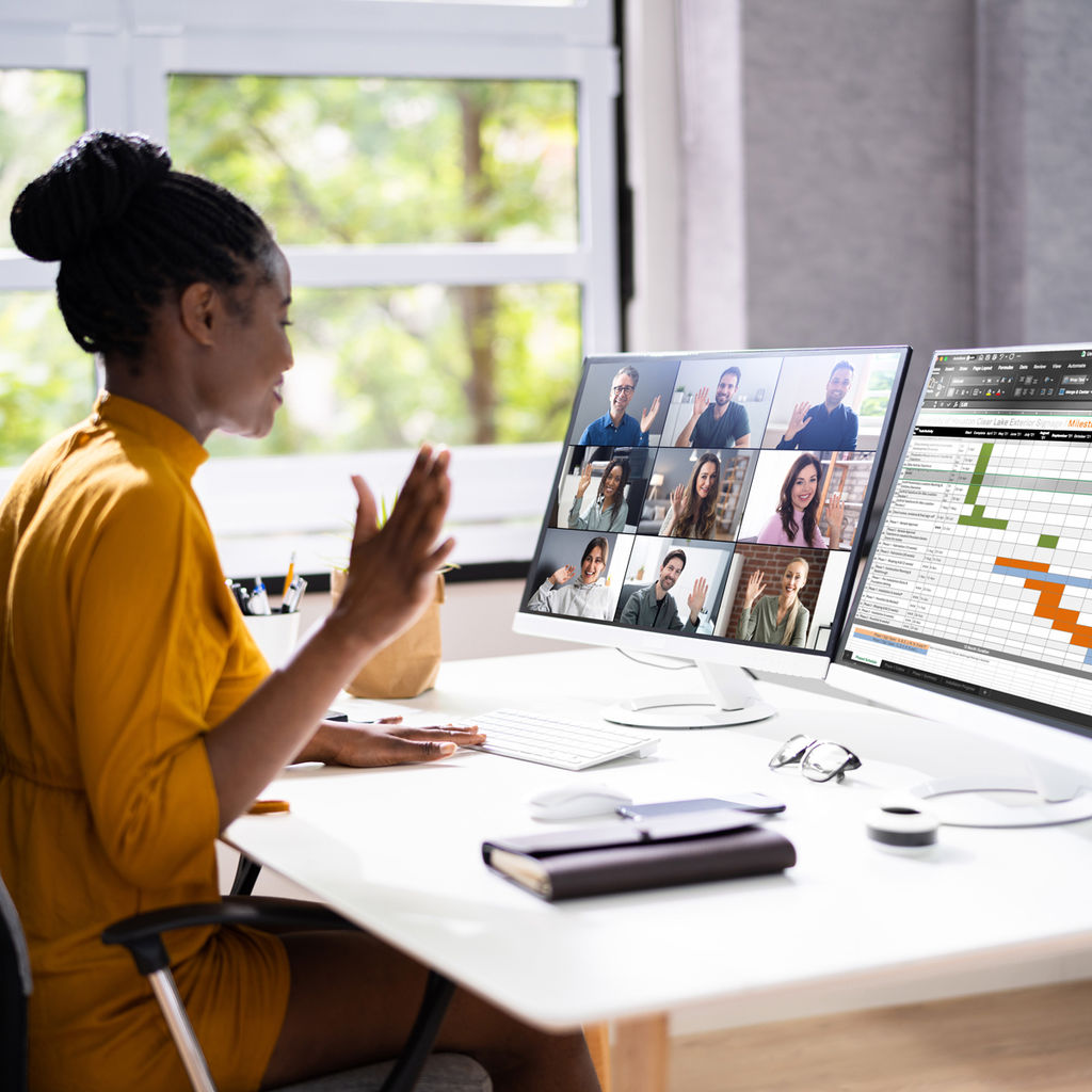 A project manager sitting in front of two monitors. One monitor showing other members of a virtual call and the other has a project schedule.