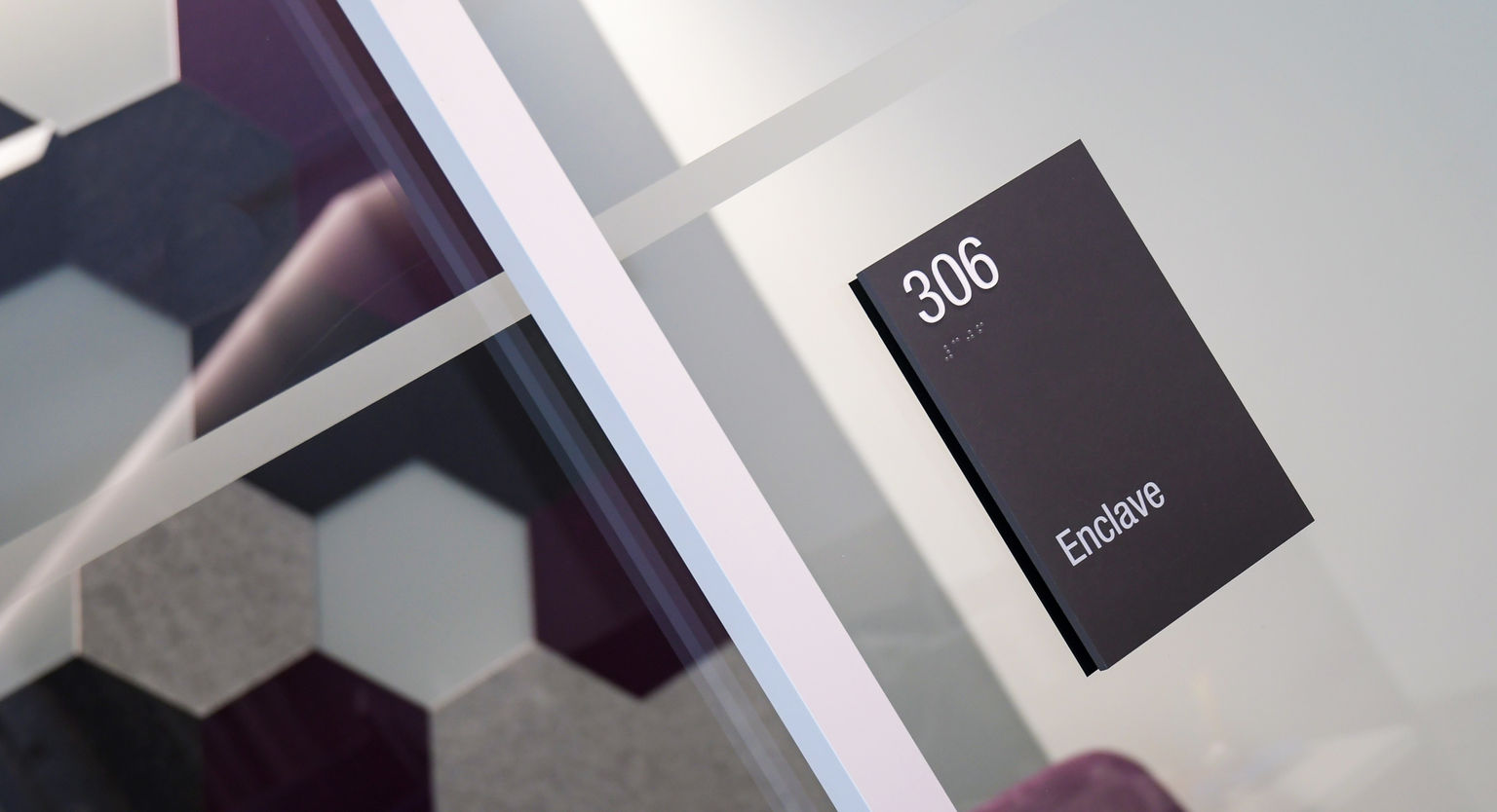 Contemporary black minimalist room identifaction sign mounted on glass office wall. Colorful acoustic wall tiles visible in background.