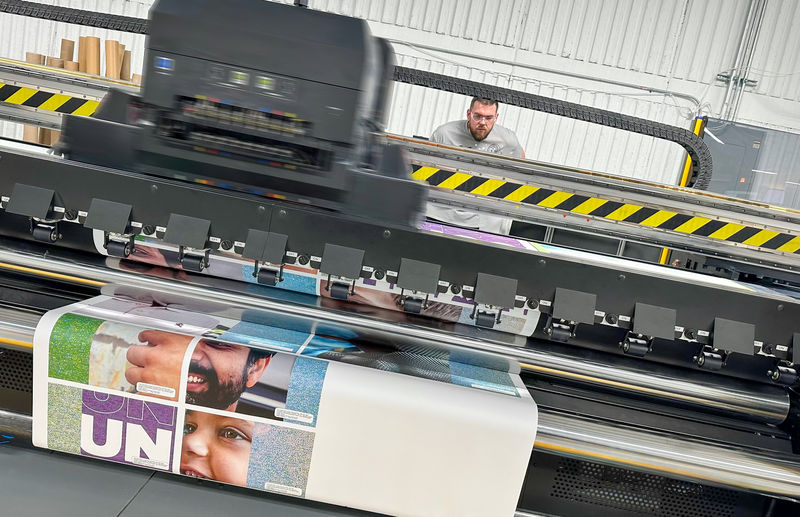 A man standing behind a large format CMYK printer running