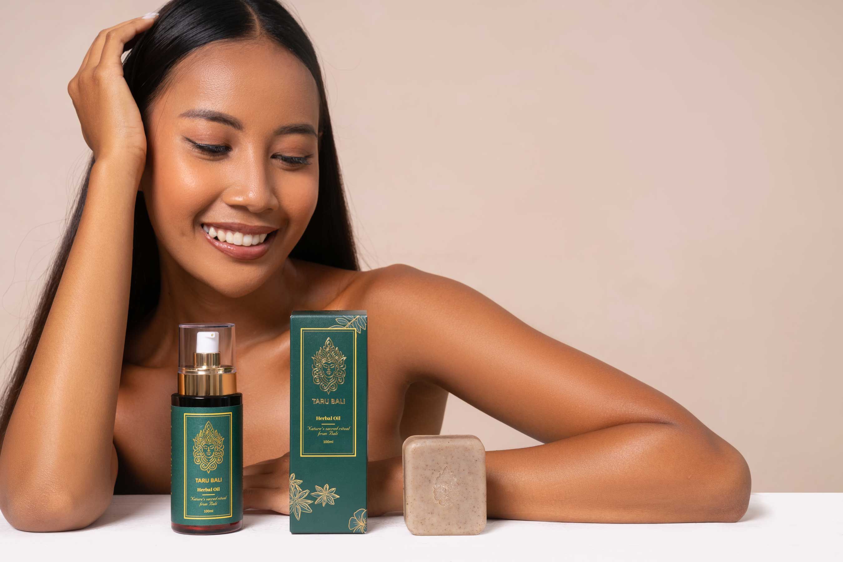 Woman with glowing healthy skin sitting behind and looking at the Taru Bali organic products - 108-herb wellness oil, luxury packaging, and natural herbal soap made from a traditional Balinese recipe