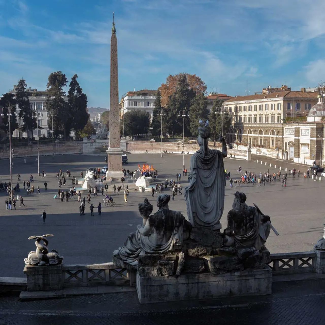 Panoramic view of Rome's Piazza del Popolo: obelisk centerpiece, statues in foreground, historic buildings around the square.