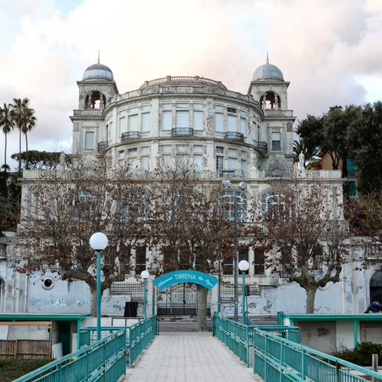 Historic white villa with domed towers and ornate facade, framed by bare trees and a turquoise pedestrian bridge.