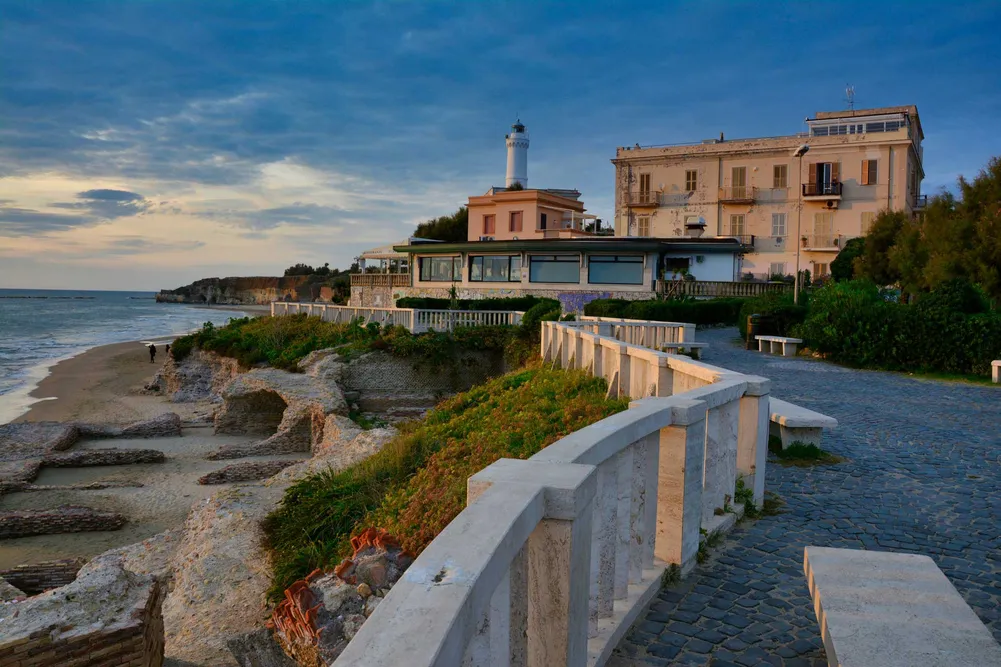 Coastal town with a lighthouse, weathered buildings, and a cobblestone promenade along the shore at sunset.