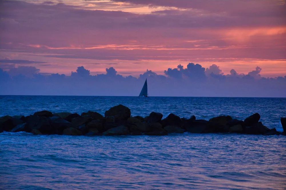 Sailboat on calm ocean at sunset with purple-pink sky and rocky foreground.