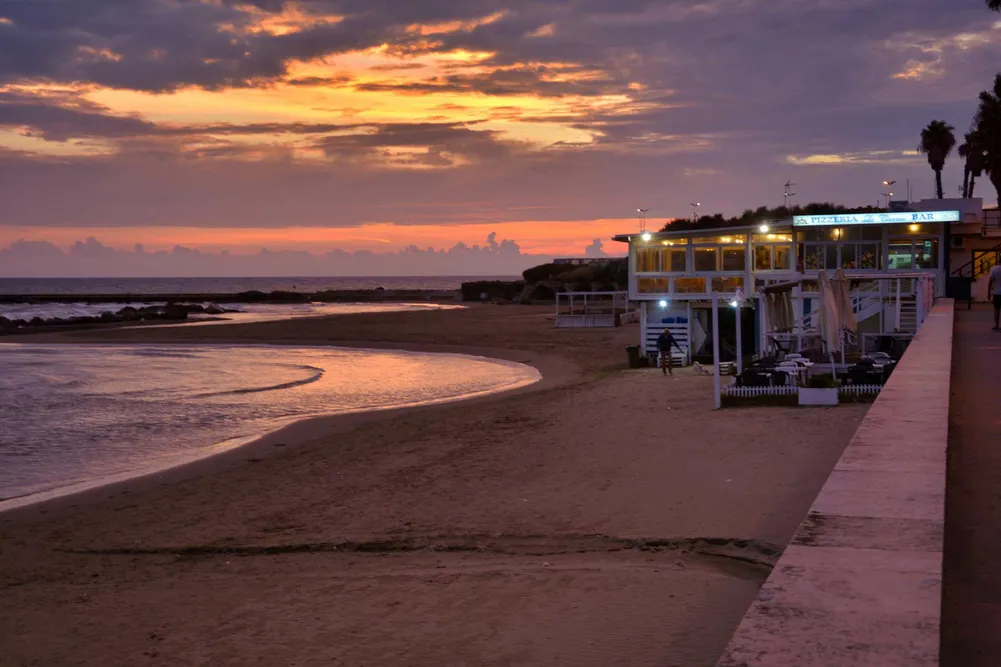 Sunset over a calm beach with a two-story seaside pizzeria and bar, palm trees and a boardwalk by the water.