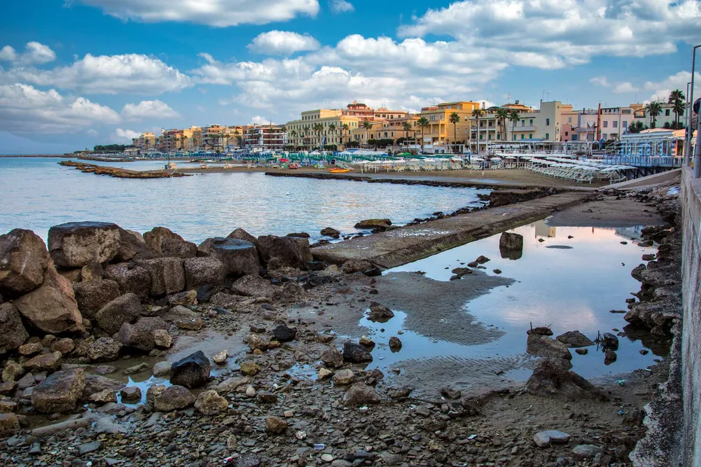 Coastal Mediterranean town with pastel buildings, palm trees, and a crowded beach with umbrellas along a rocky shore.