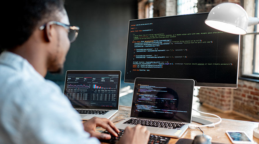 Young african male programmer writing program code sitting at the workplace with three monitors in the office. Image focused on the screen