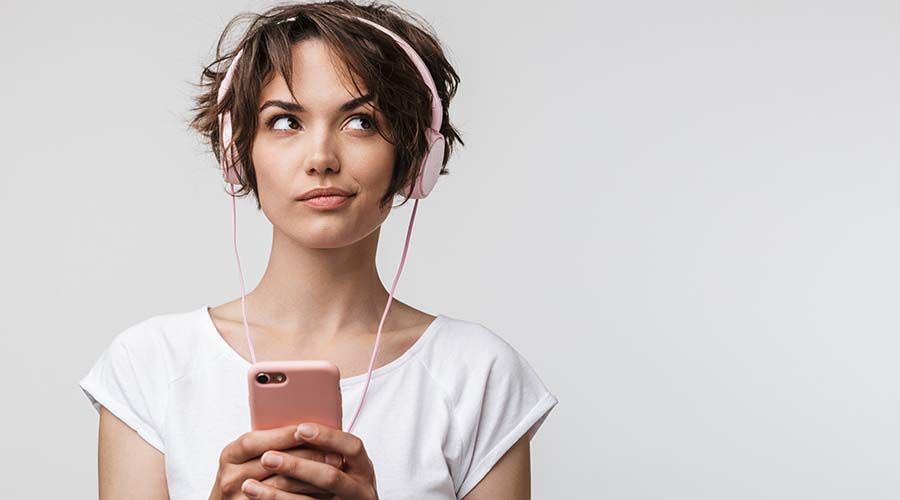 Image of joyful woman in basic t-shirt holding smartphone while listening to music with headphones isolated over white background