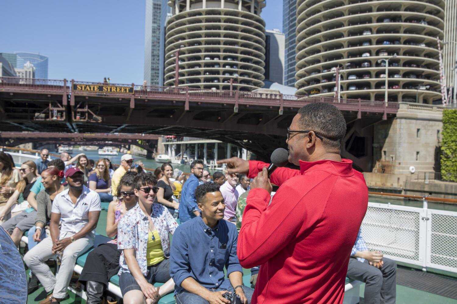 A man in a red shirt pointing and speaking into a microphone while a large group looks on. 