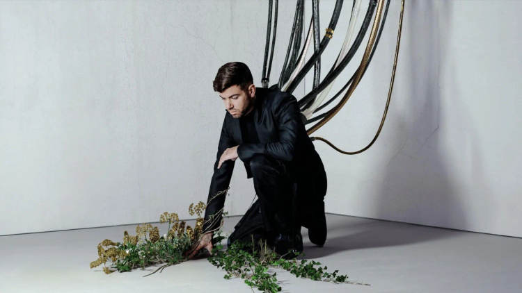 Man in dark jacket crouches beside wilted plants on white floor in gallery space with coiled cables on wall behind him Man in dark jacket crouches beside wilted plants on white floor in gallery space with coiled cables on wall behind him