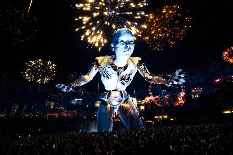 A massive illuminated figure projection towers above a nighttime crowd with fireworks bursting overhead A massive illuminated figure projection towers above a nighttime crowd with fireworks bursting overhead