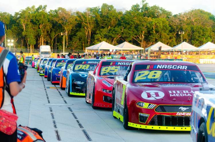 Autos de carreras de NASCAR alineados en un evento de deportes de motor, con carpas y espectadores visibles al fondo
