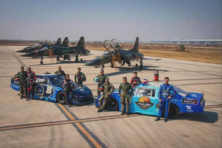 Personal militar y pilotos de carreras posan junto a coches de carreras azules en la pista de un aeropuerto, con aviones de combate estacionados al fondo.