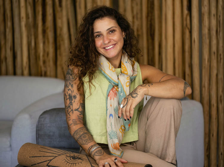 Woman with long curly dark hair and multiple tattoos sitting on a gray couch against a gold curtain backdrop, wearing a yellow top and patterned scarf Woman with long curly dark hair and multiple tattoos sitting on a gray couch against a gold curtain backdrop, wearing a yellow top and patterned scarf