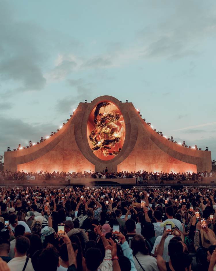 Large crowd of spectators holds up phones while watching a massive stage structure with an illuminated oval screen displaying imagery against a cloudy sky at du Large crowd of spectators holds up phones while watching a massive stage structure with an illuminated oval screen displaying imagery against a cloudy sky at du