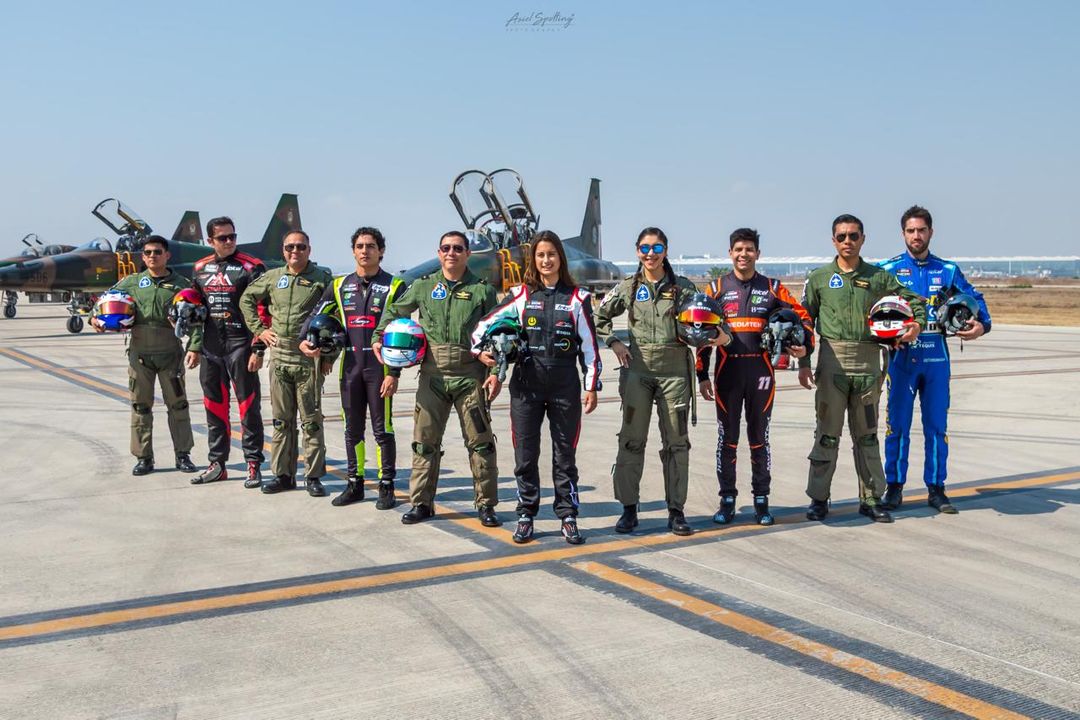 Grupo de pilotos y personal de aviación con trajes de vuelo de pie en la pista de un aeropuerto con un avión militar al fondo.