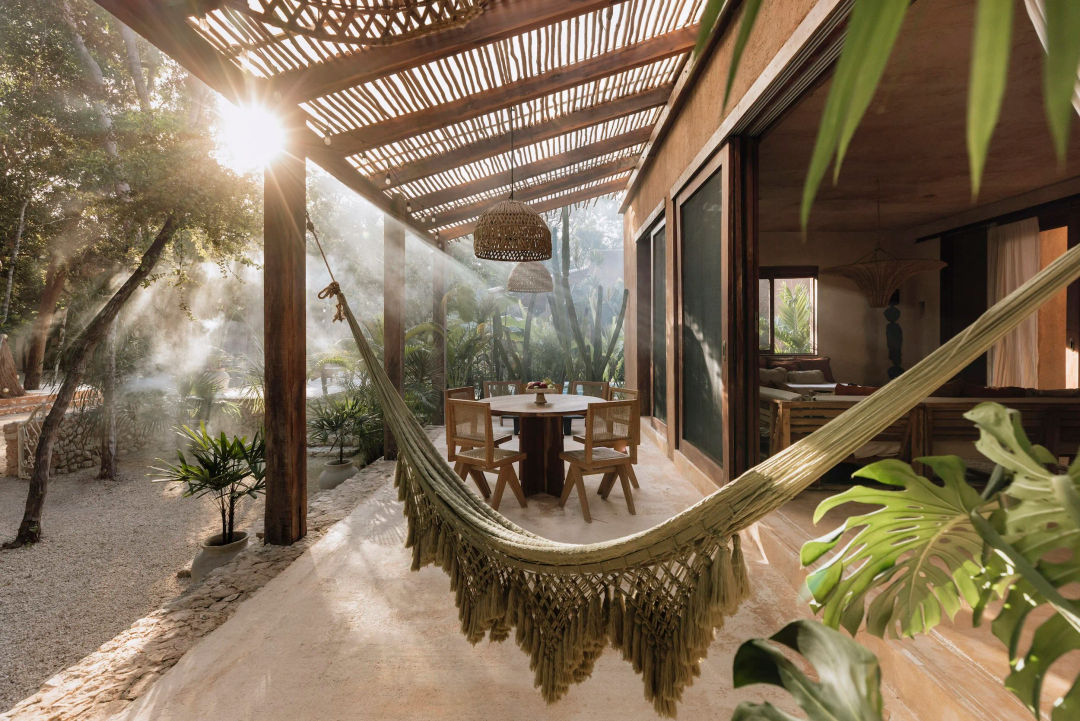Una hamaca cuelga en un patio techado junto a una mesa de comedor, con plantas tropicales y palmeras de fondo bajo la luz que se filtra entre las hojas