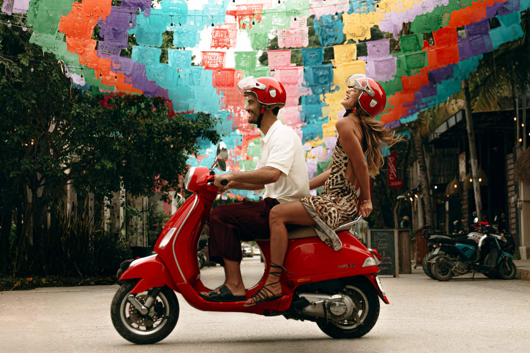 Red against Caribbean blue, the campaign that photographs Tulum as one single world Two people wearing red helmets ride a red scooter down a narrow street beneath a colorful overhead canopy of paper flags