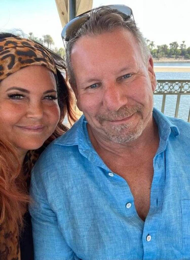Two people pose for a selfie on a waterfront deck with palm trees and water visible in the background Two people pose for a selfie on a waterfront deck with palm trees and water visible in the background