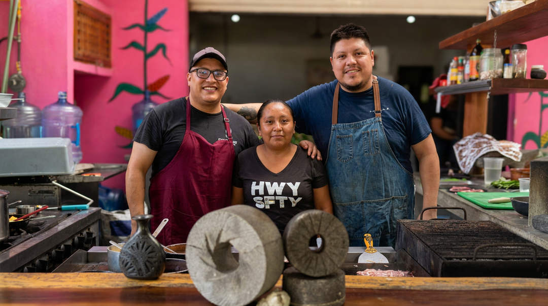 The most incredible meal near Tulum is authentic, slow, ancient and delicious Three people stand behind a kitchen counter with large traditional grinding stones in the foreground, with pink walls and cooking equipment visible in the backg
