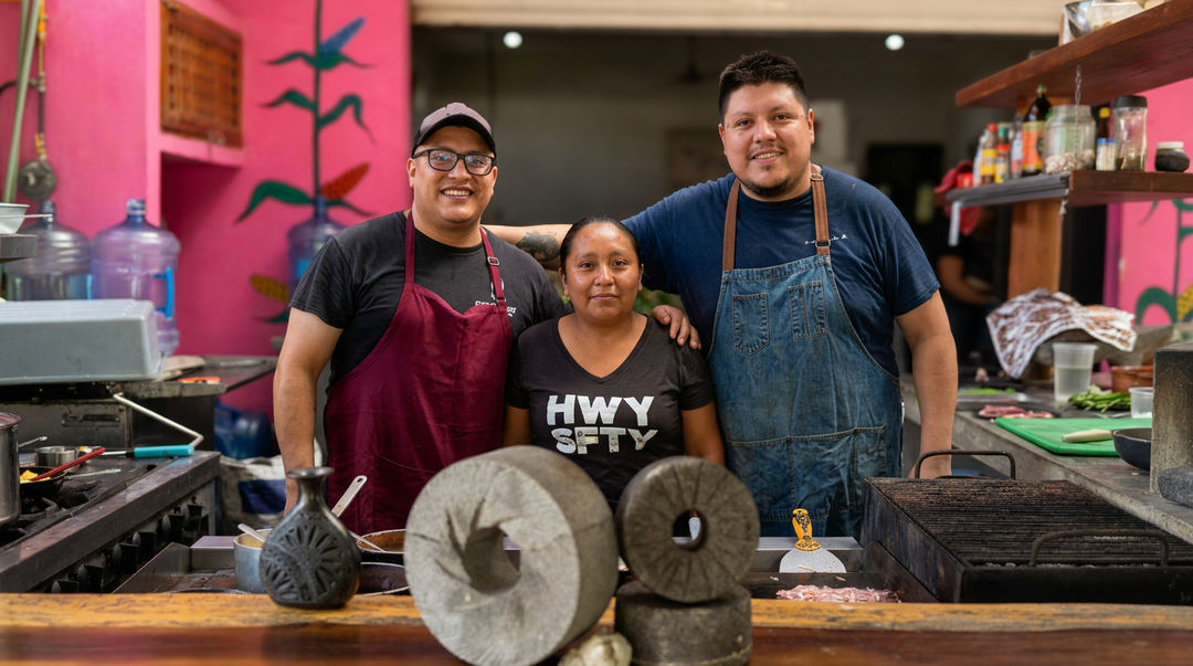The most incredible meal near Tulum is authentic, slow, ancient and delicious Three people stand behind a kitchen counter with large traditional grinding stones in the foreground, with pink walls and cooking equipment visible in the backg