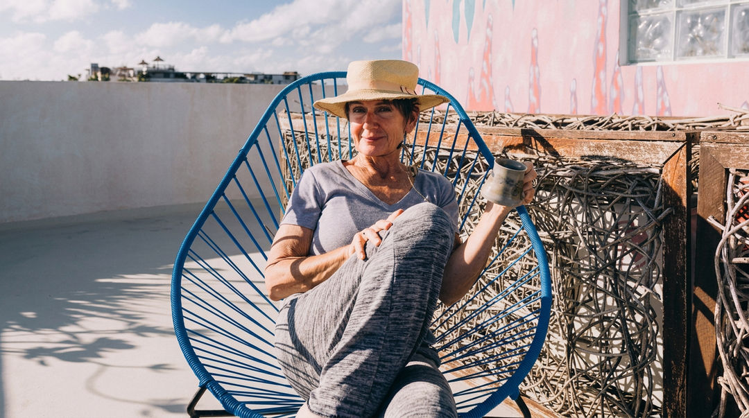 Fanny Barry Came to Tulum to Heal and Stayed to Write a Superhero Trilogy Woman in a straw hat sits in a blue woven chair on a rooftop surrounded by fishing nets and a pink building