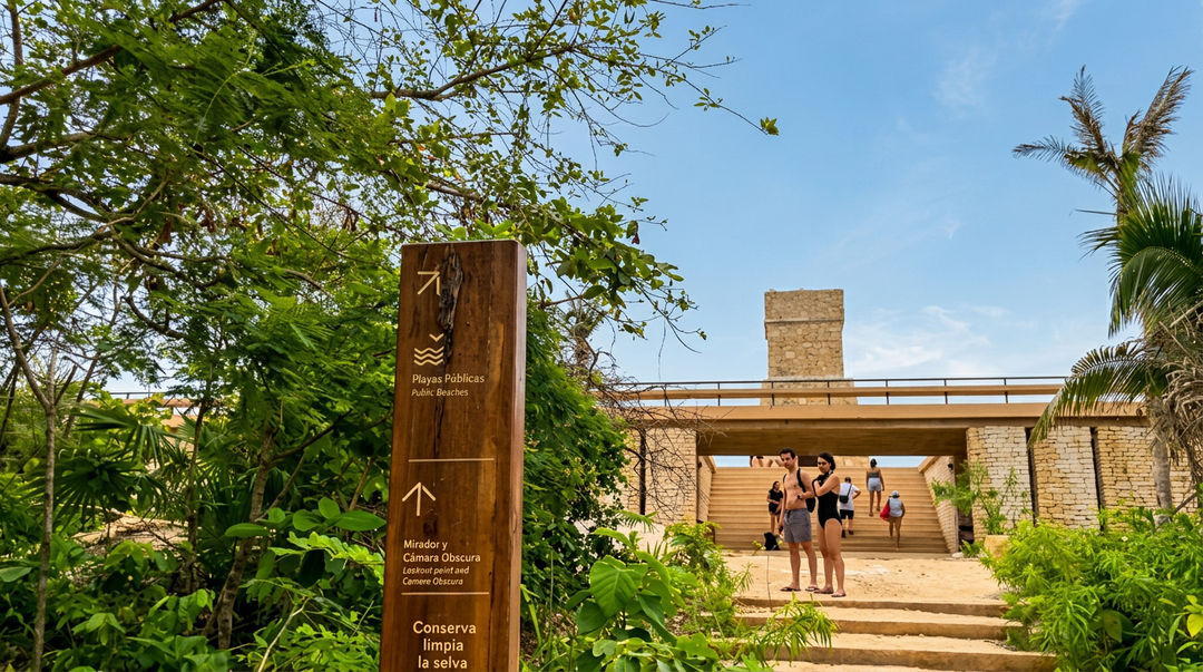 Señalamiento de madera entre vegetación tropical guía a los visitantes hacia edificio moderno de ladrillo con techumbre