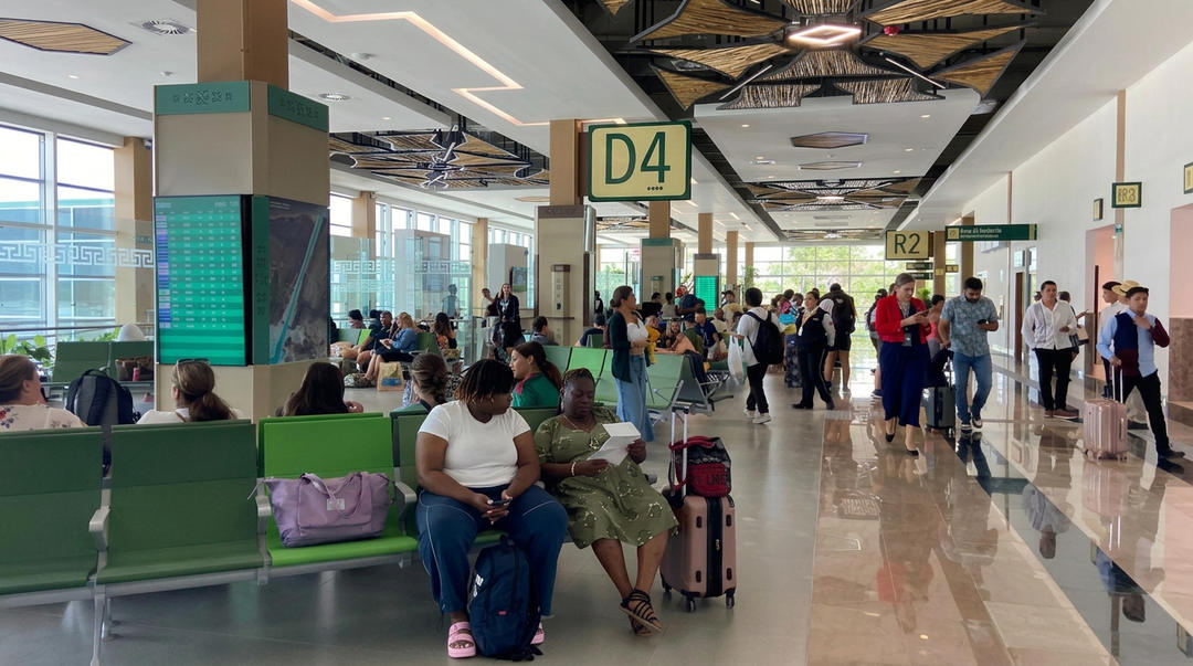 Passengers wait and move through a busy airport terminal with green seating areas and directional signage overhead