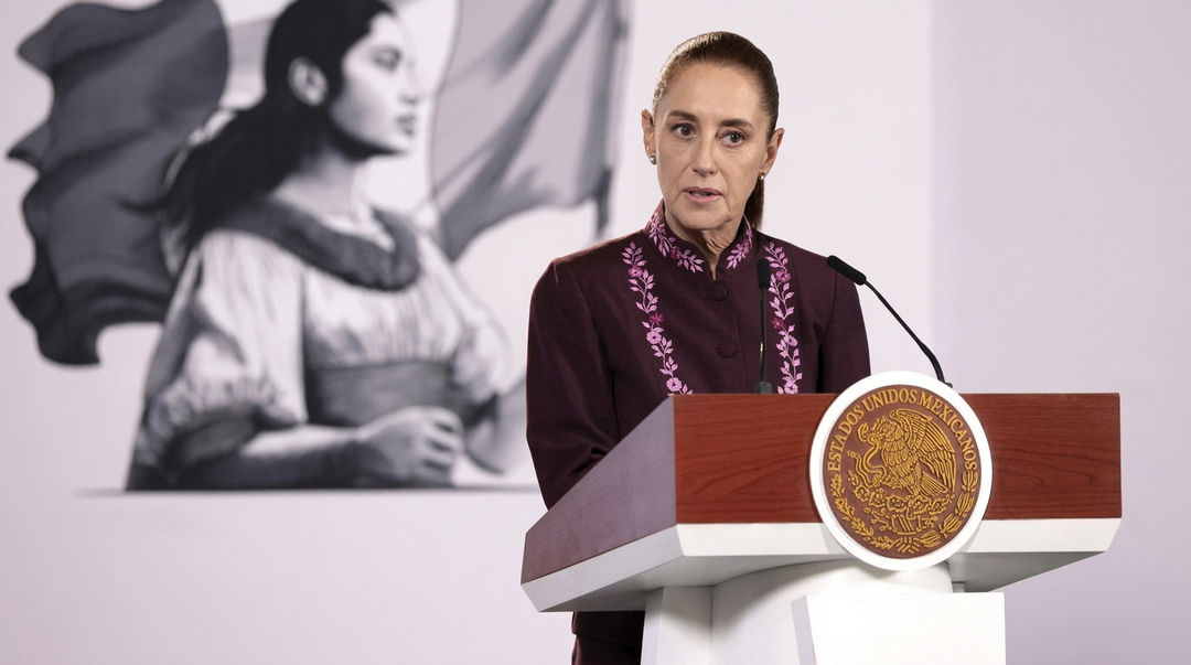 Sheinbaum answers the security question behind Tulum flights Woman in maroon embroidered shirt speaking at a podium with an official seal, with a mural of a figure visible in the background