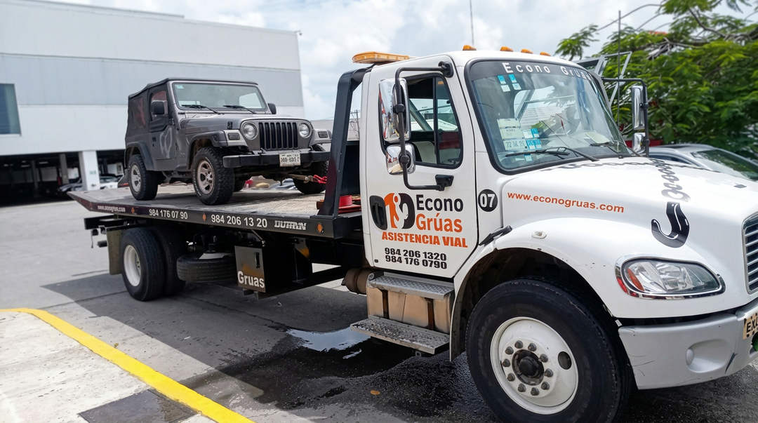 A white tow truck carries a dark-colored jeep on its flatbed in a parking lot