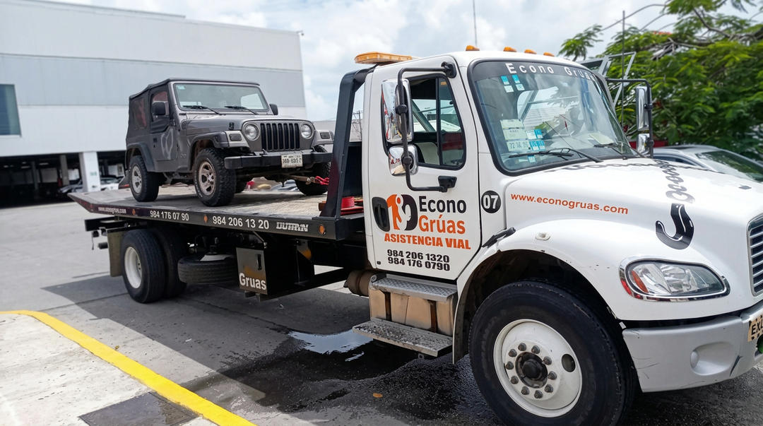 A white tow truck carries a dark-colored jeep on its flatbed in a parking lot