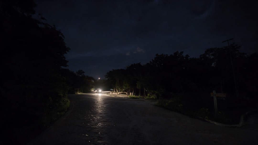 Por qué las calles de Tulum se están quedando a oscuras A dimly lit street lined with trees shows sparse streetlights illuminating a mostly dark roadway at night