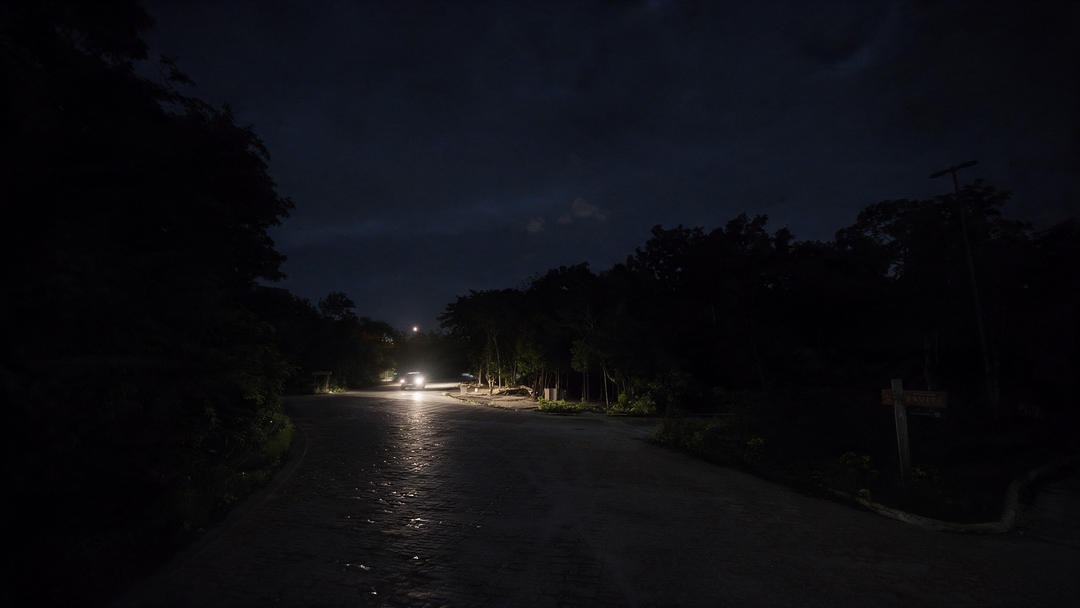 A dimly lit street lined with trees shows sparse streetlights illuminating a mostly dark roadway at night