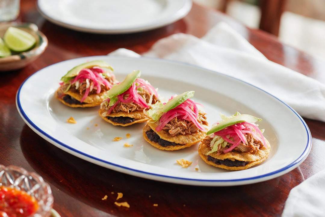 Four fried tortillas stuffed with black bean paste, topped with shredded pork, pickled red onion and avocado, on a cobalt rimmed platter