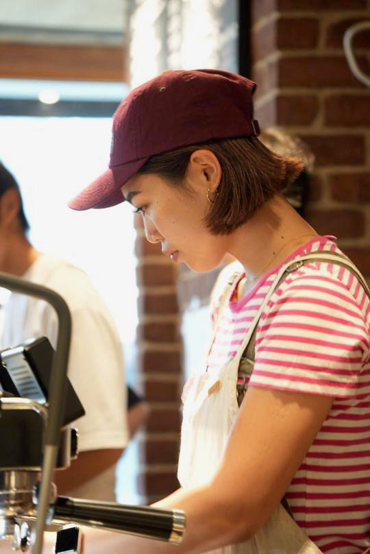 yumi in an apron and a cap shot from the left side of her crafting coffee