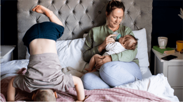 Mother sat in bed feeding a baby milk from an Advanced anti-colic bottle. Another child is doing a headstand on the bed