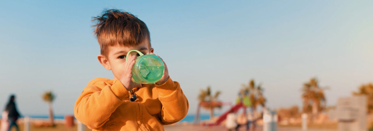 Toddler drinking from sportee water bottle whilst sat outside in front of a beach