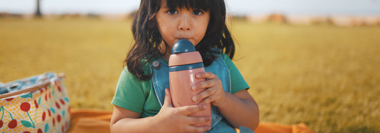 Toddler drinking from insulated straw cup whilst sat outside on a field 