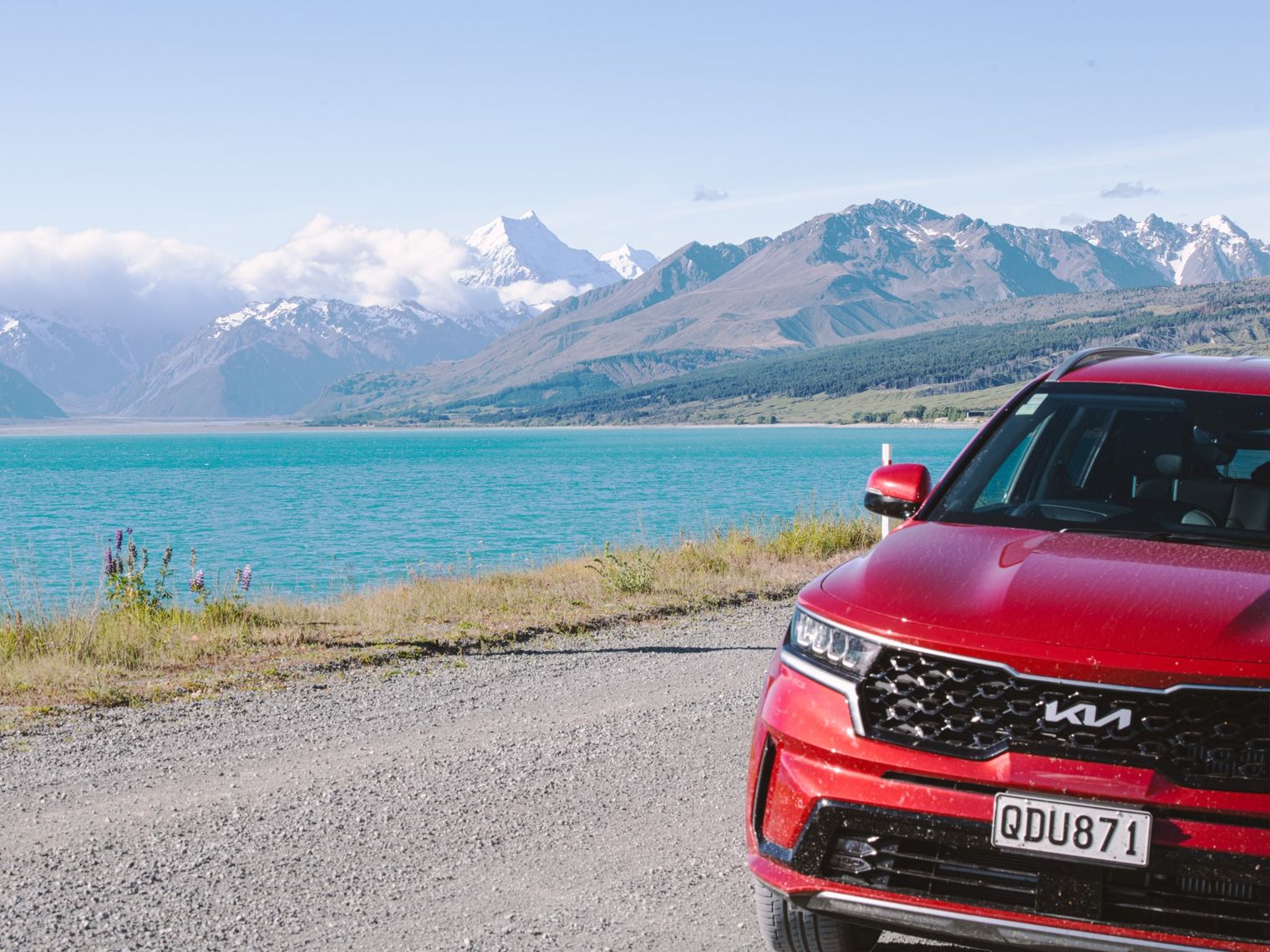 A red car parked up on a gravel road next to a blue lake with snowy mountains in the background